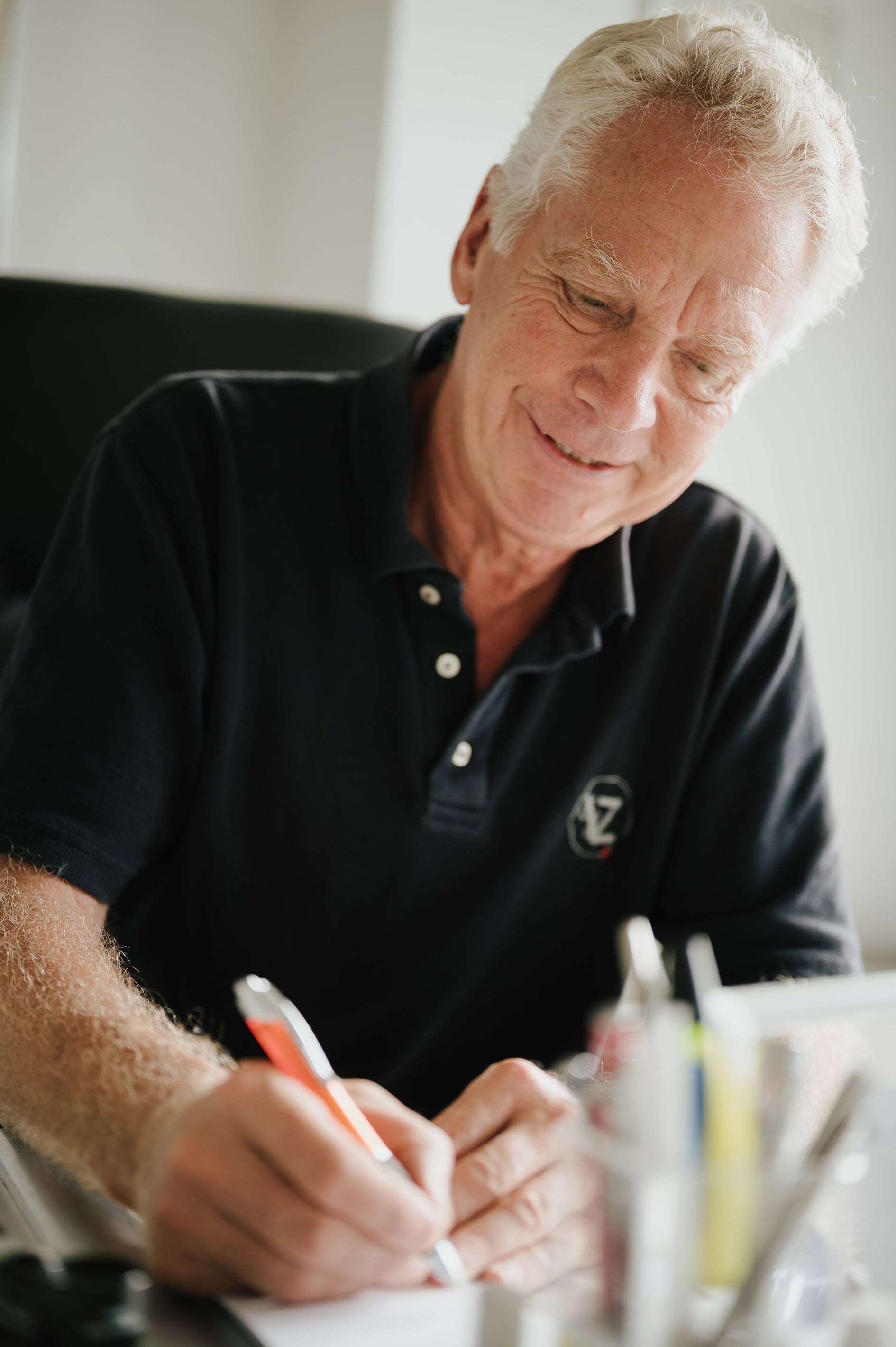 Dr. Volker Zähres schreibt etwas, schaut nach unten und lächelt freundlich, als würde er im Gespräch mit Patient:innen etwas mitschreiben. Er trägt ein schwärzes Polo-Shirt mit seinen Initalien.